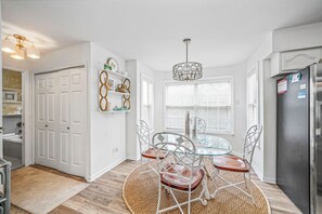 Een moderne keuken met witte kasten, een roestvrijstalen koelkast, een glazen eettafel met metalen stoelen en een decoratieve hanglamp.