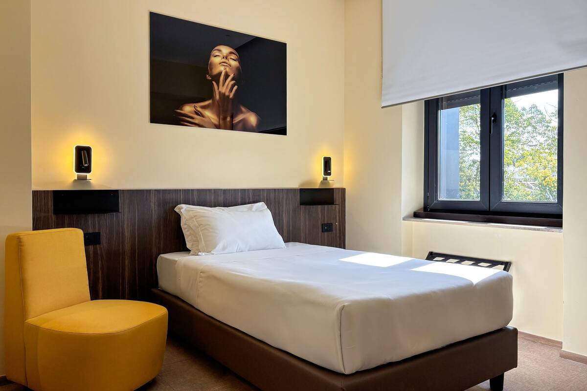 A hotel room with a bed, a bedside table, a chair, a window, and a wall-mounted picture.