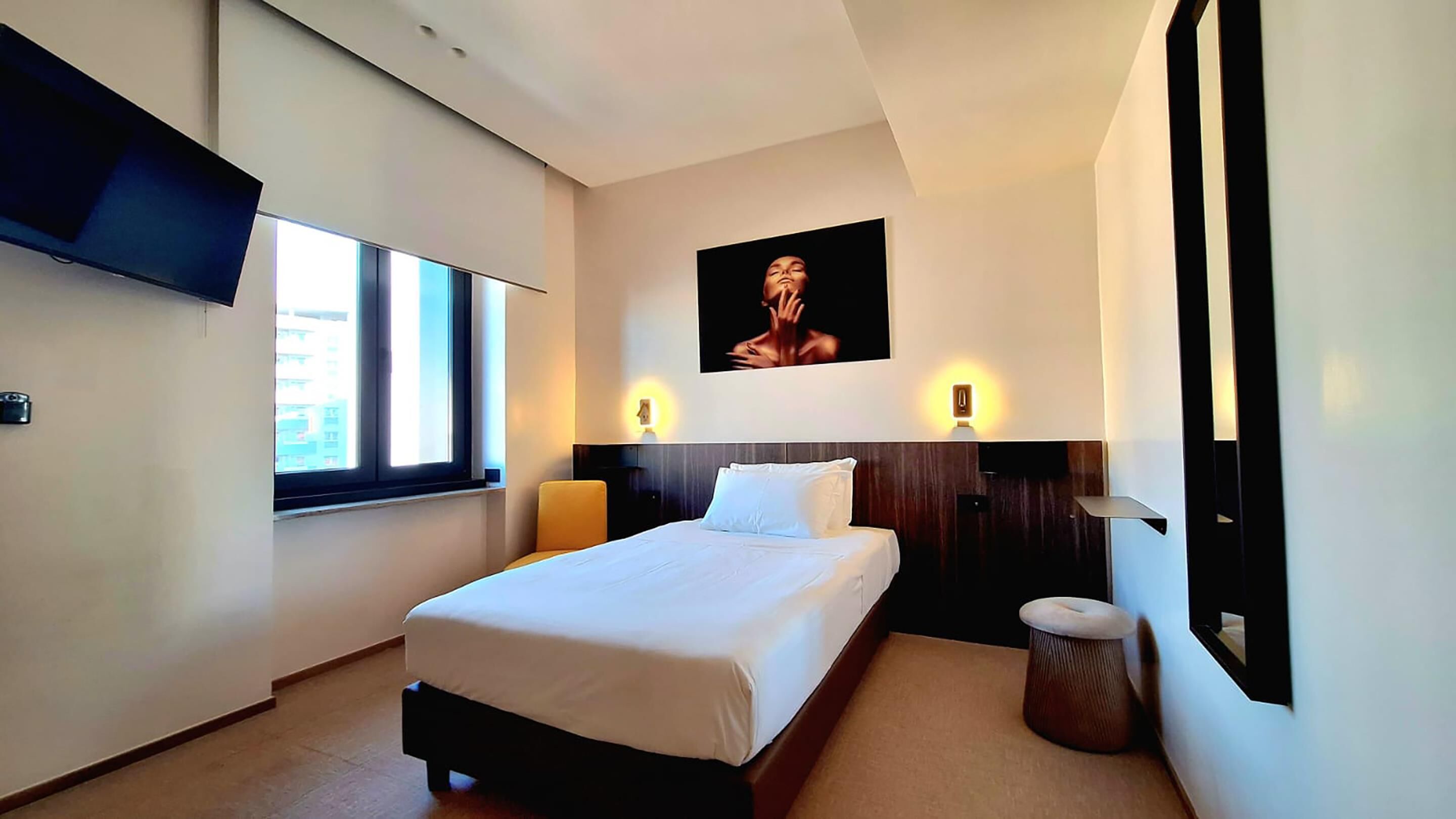 Ein Hotelzimmer mit Bett, einem an der Wand montierten Fernseher, einem Fenster und einem Bild.