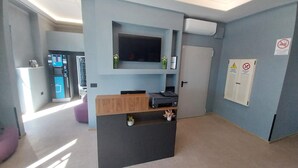 Una habitación moderna con una televisión de pantalla plana, un escritorio con impresora, una puerta con señal de advertencia y una máquina expendedora.