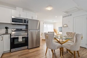 Una cocina moderna con electrodomésticos de acero inoxidable, un comedor con mesa de vidrio y sillas tapizadas, y una puerta blanca.