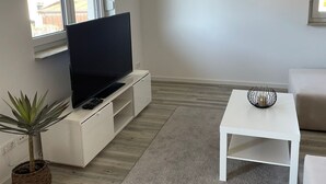 A modern living room with a flat-screen TV on a white cabinet, a small white table with a bowl, a potted plant, and a grey rug. - Newly built apartment with lake view-Teupitzer Str.9 Tornow (Teupitz)