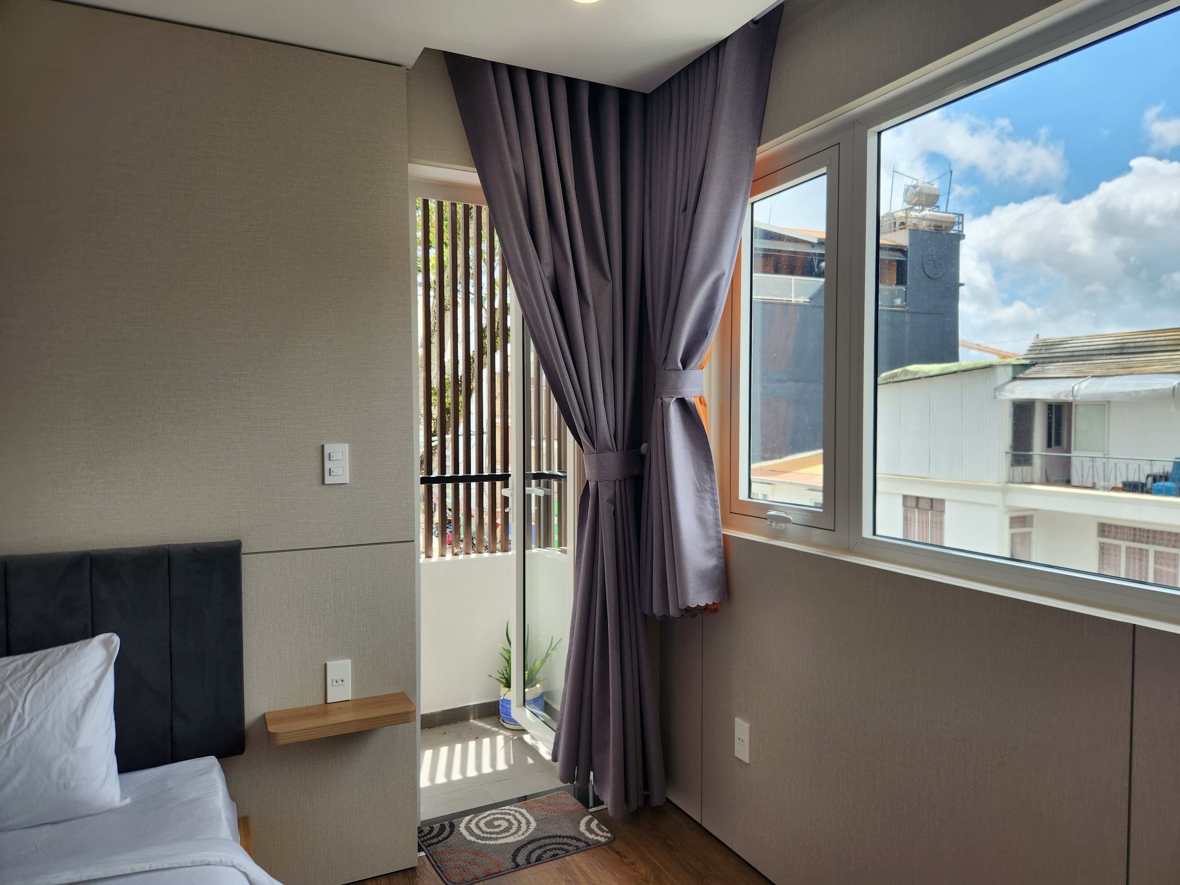 A hotel room with a bed, a window with curtains, a small plant, and a view of buildings outside.