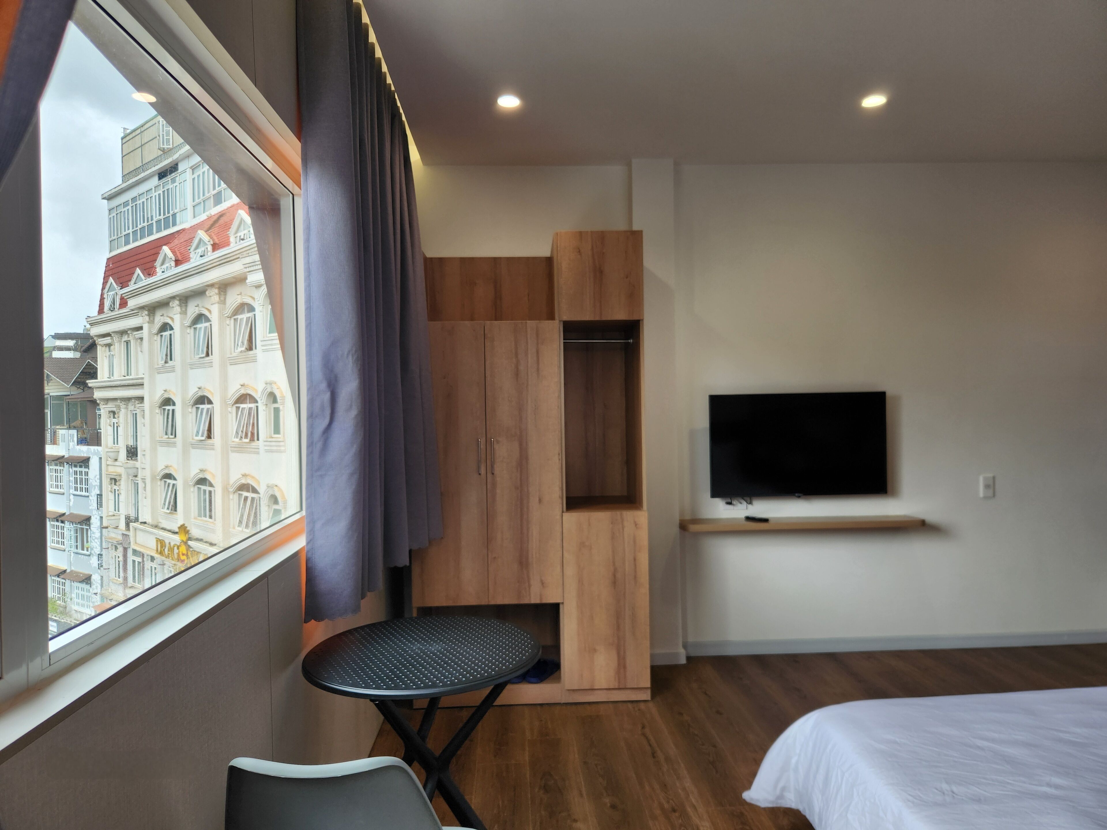 A modern hotel room with a large window, a bed, a TV mounted on the wall, and a wooden wardrobe.