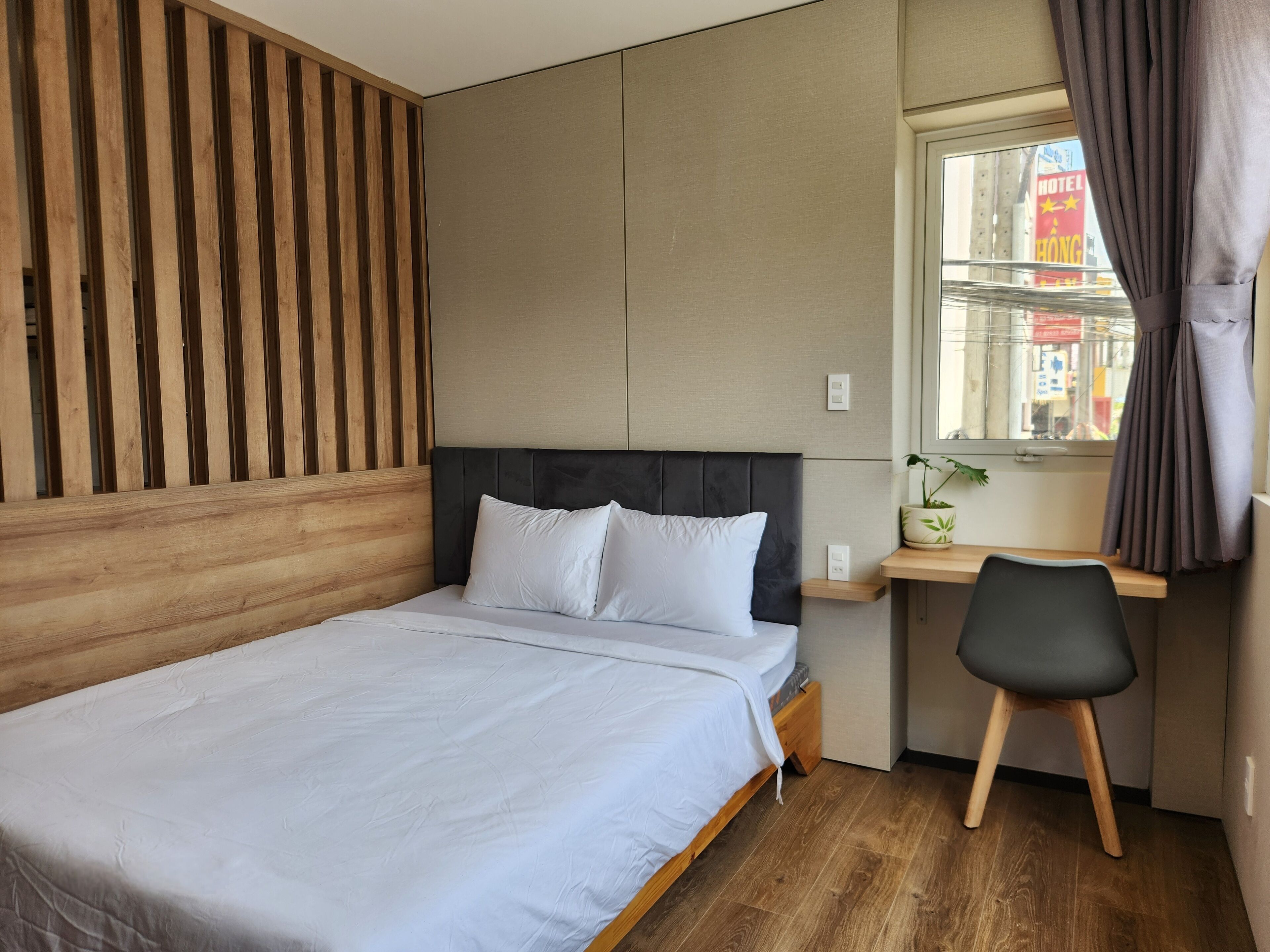 Ein ordentlich bezogenes Bett mit weißen Bettwäsche, eine Wand aus Holzleisten, ein kleiner Schreibtisch mit Stuhl und ein Fenster mit Vorhängen.