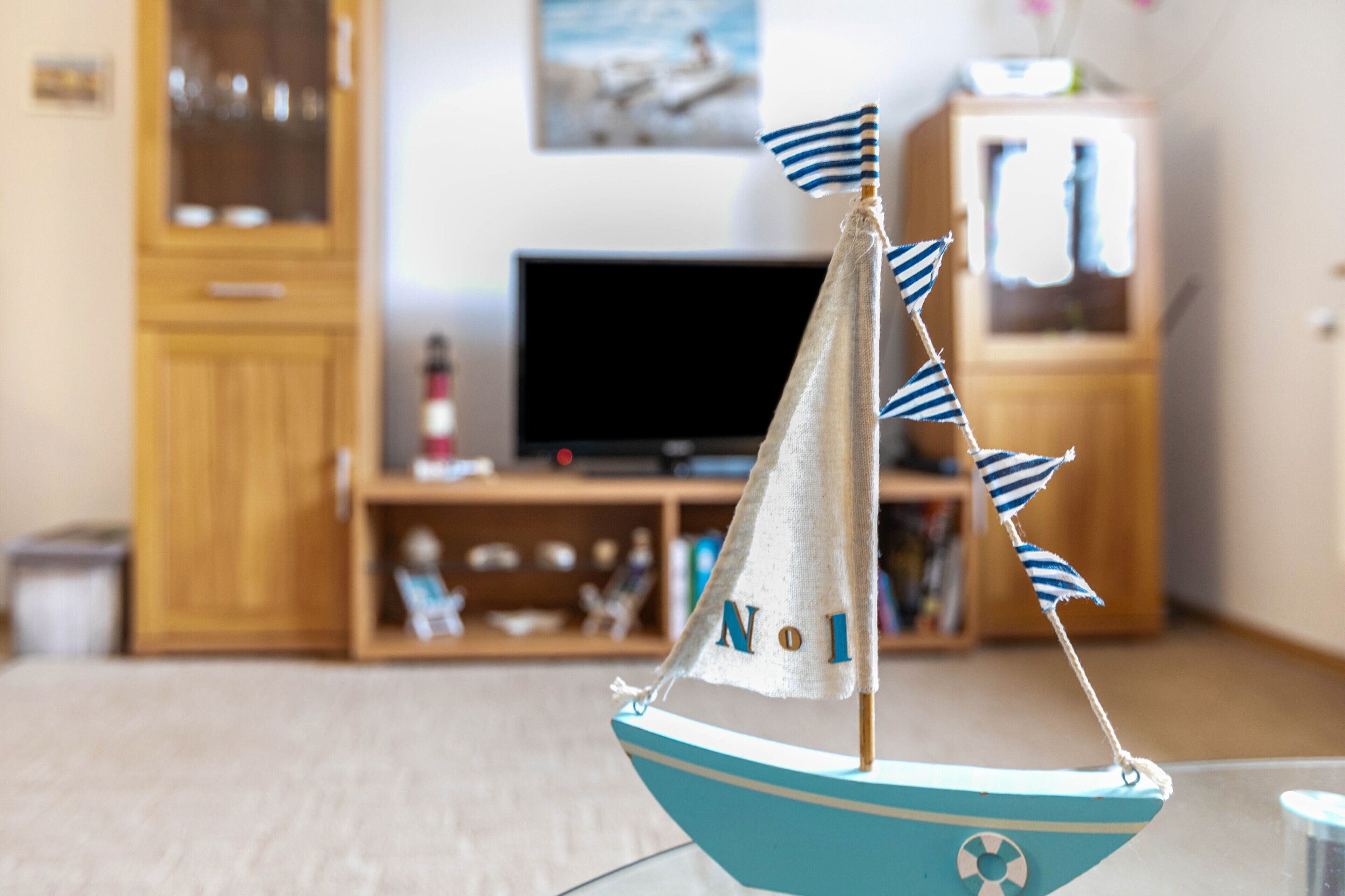 Ein Modellsegelboot mit den Buchstaben 'NO' darauf, platziert in einem Zimmer mit einem Holzkabinett, einem Fernseher und einem Regal mit verschiedenen Gegenständen.