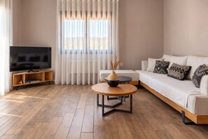 A modern living room with a sofa, a coffee table, a TV, and a window with sheer curtains. - Cozy, family-friendly apartment with a view! (Skiathos)
