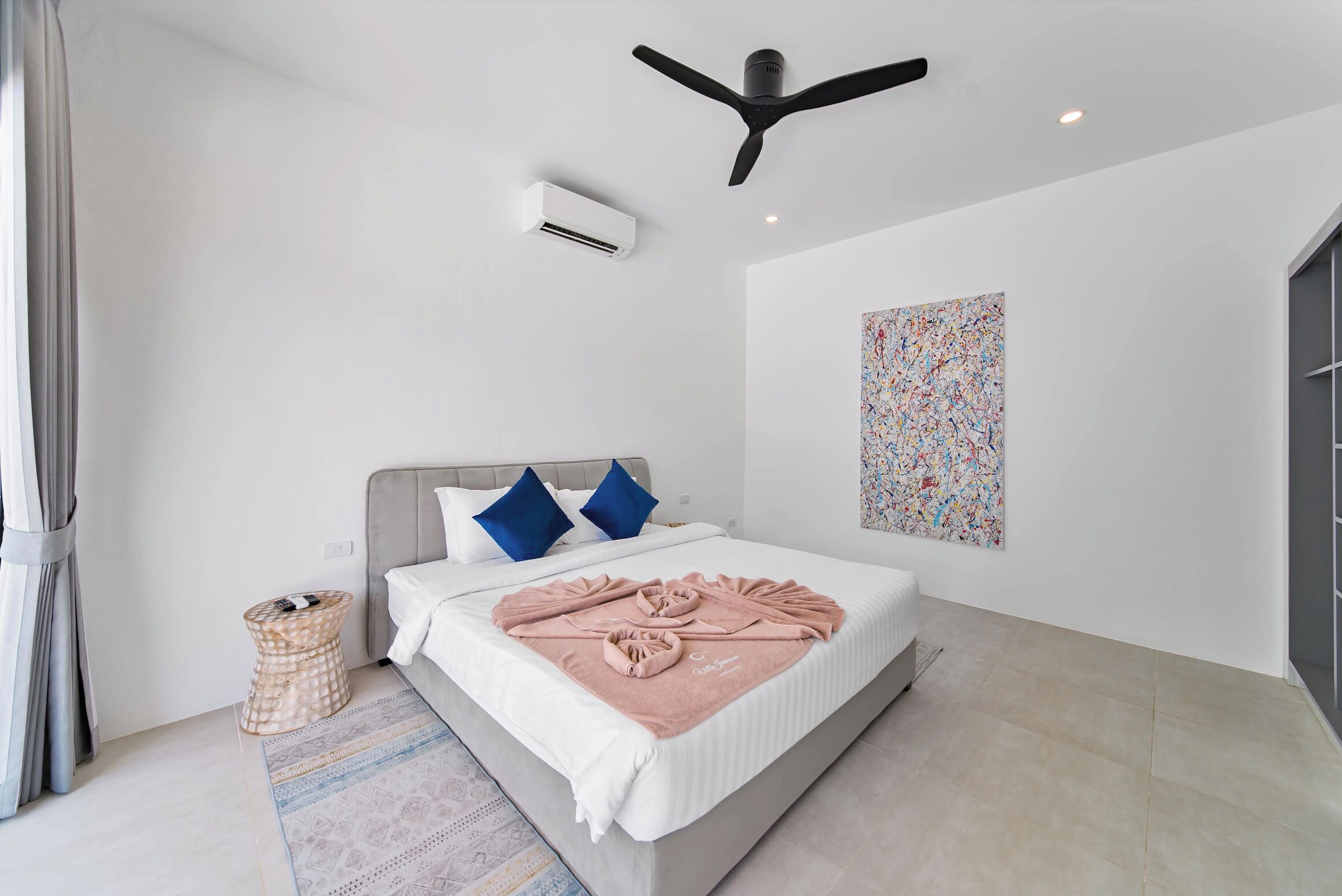 A modern hotel room with a bed, air conditioning unit, ceiling fan, and a colorful abstract painting on the wall.