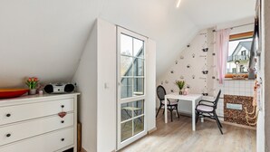 Quarto com penteadeira branca, mesa com cadeiras, janela e vista para uma casa lá fora.
