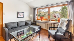 Uma sala de estar com sofá, poltrona, mesa de centro de vidro, janela com cortinas e radiador.