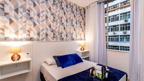 Ein Hotelzimmer mit Wand im geometrischen Muster, einem Bett mit blauen Kissen, einem Nachttisch mit Lampe und einem Fenster mit Vorhängen.