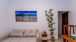 Ein Wohnzimmer mit einem beigefarbenen Sofa, einem Holzesstisch und einer Topfpflanze. Drei an der Wand hängende Bilder zeigen eine Strandansicht.
