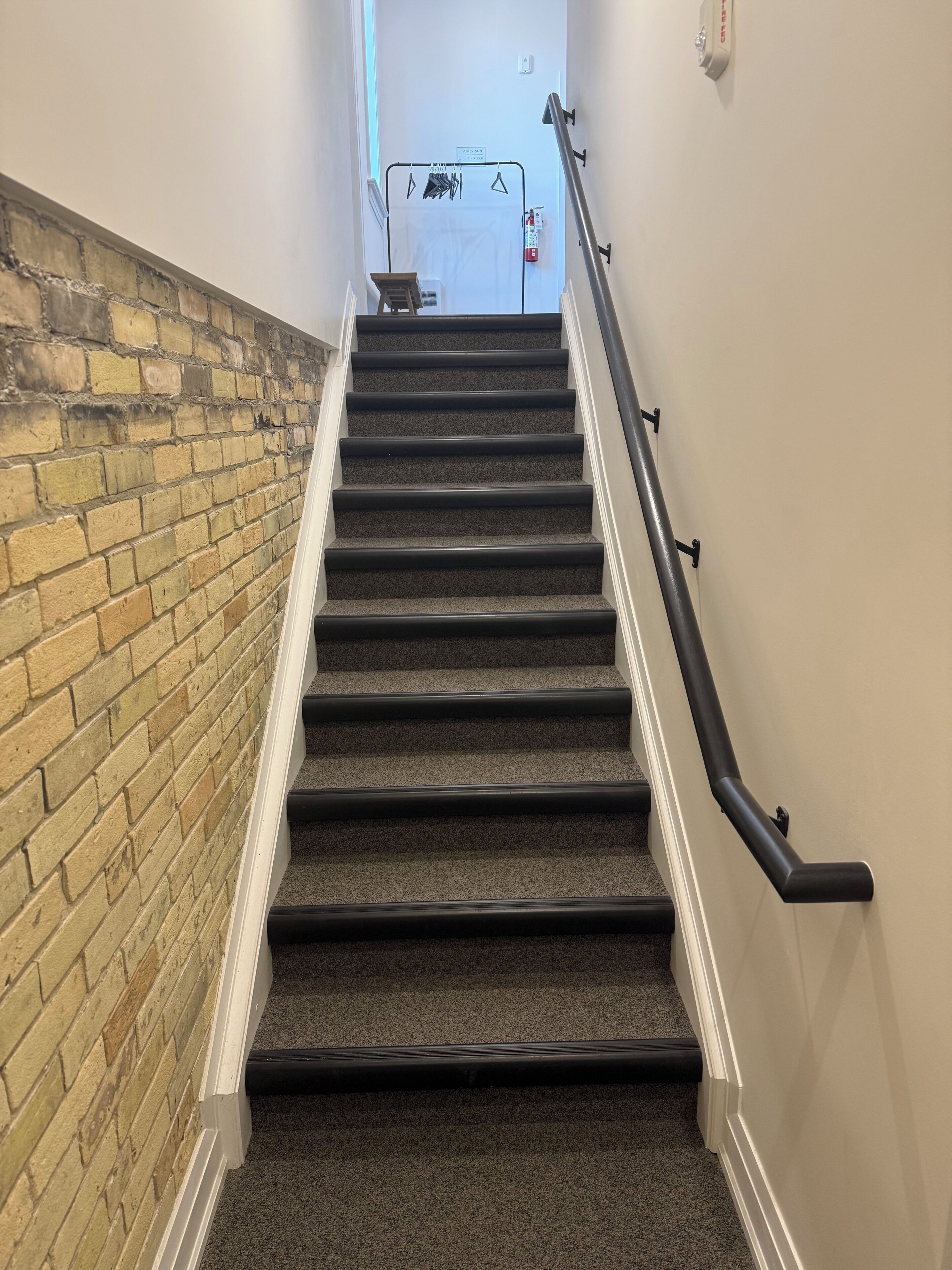 A staircase with carpeted steps and a handrail, leading to a room with a white door and a fire extinguisher mounted on the wall.