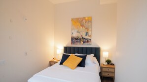 A neatly made bed with a white duvet, a dark headboard, and a yellow pillow. There are two bedside tables with lamps and a clock. A colorful abstract painting hangs above the bed.