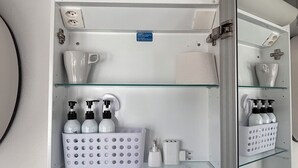 Casa de banho com estantes brancas, um espelho e vários artigos de higiene pessoal.