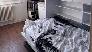Quarto com cama, televisão na parede, radiador e janela com vista para o verde lá fora.