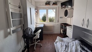 Quarto compacto com cama, escrivaninha e cadeira. Janela com vista para o verde lá fora.