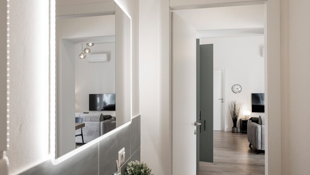 A modern interior with a hallway leading to a bedroom. A large mirror reflects a TV, a bed, and a nightstand. The room has a wooden floor and a white door. - [Leopoldo Cozy Loft] Tram| Air C. | Smart TV (Firenze)
