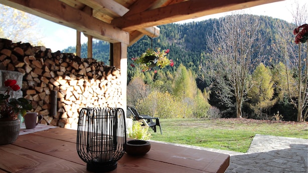 Una mesa de madera con una cesta de alambre negra y un cuenco, un muro de piedra con leña apilada, y vista a una ladera boscosa.