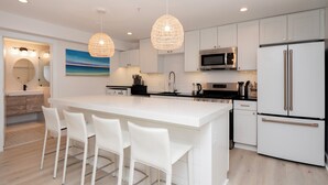 A modern kitchen with white cabinetry, a large island with bar stools, stainless steel appliances, and a hanging light fixture. - War College Friendly Downtown Condo (Newport)