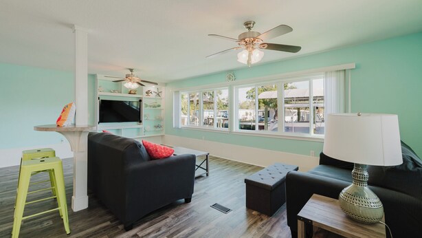A living room with a sofa, ottoman, and a small table with a lamp. There is a TV mounted on the wall and a ceiling fan. - Waterfront Escape • Private Dock • Near 3 Sisters Springs (Crystal River)