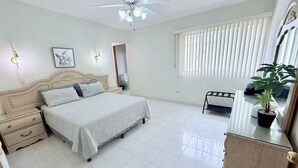 A bedroom with a bed, bedside tables, a ceiling fan, a window with blinds, and a potted plant.