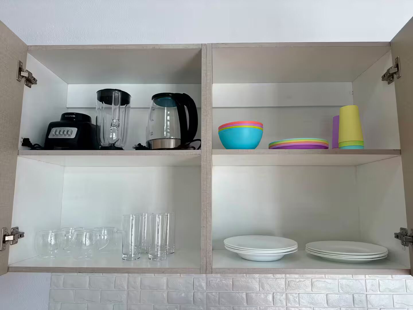 A kitchen with a white tiled backsplash, two shelves with kitchen appliances, and various dishes and glasses.