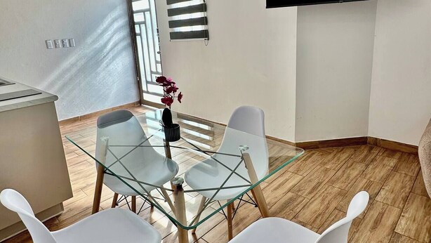 A modern living room with a glass dining table, white chairs, a flat-screen TV mounted on the wall, and a window with blinds. - Very Central | Tec Mty | Hosp Angeles | Invoicing (Torreón)