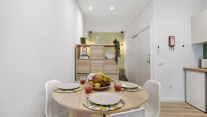 Un espace repas compact avec une table ronde, des chaises blanches, un panier de fruits et un petit coin cuisine équipé d’un lave-vaisselle.