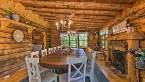 A rustic wooden cabin interior with a large round dining table surrounded by white chairs, a stone fireplace, and a ceiling fan.