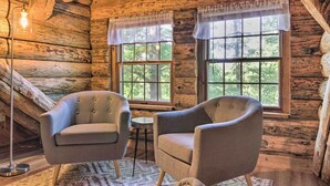 A cozy log cabin room with two upholstered chairs, a small round table, and a floor lamp. Large windows with curtains provide natural light and views of the surrounding forest.