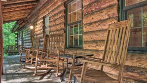 A log cabin with a porch featuring wooden rocking chairs and tables.