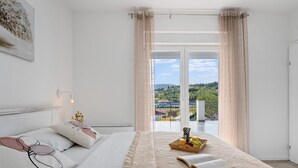A bedroom with a bed, a bedside table with a lamp, a window with curtains, and a view of greenery outside. - Villa Gloria: Stylish 5-Bedroom Retreat with Private Pool and Stunning Valley Views (Zmijavci)