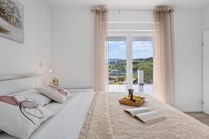 Ett sovrum med en säng, ett sängbord med en lampa, ett fönster med gardiner och utsikt över grönområden utanför.