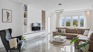 A modern living room with a flat-screen TV, a sofa, a coffee table, and a large window offering a view of the outdoors. - Villa Gloria: Stylish 5-Bedroom Retreat with Private Pool and Stunning Valley Views (Zmijavci)