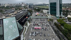 Un bâtiment moderne doté d’une façade en verre, un parking avec plusieurs rangées de voitures et un grand espace vert arboré.
