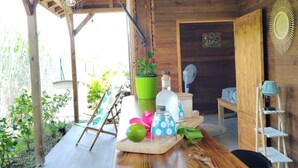 Une cabane en bois avec une terrasse en bois, une petite table sur laquelle est posée une plante en pot, un ventilateur et une horloge au mur.