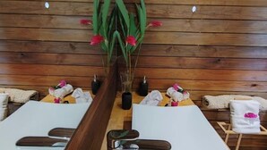 Une salle de spa aux murs lambrissés, avec une plante centrale aux fleurs rouges et deux tables de massage, équipées de serviettes et de bouteilles.