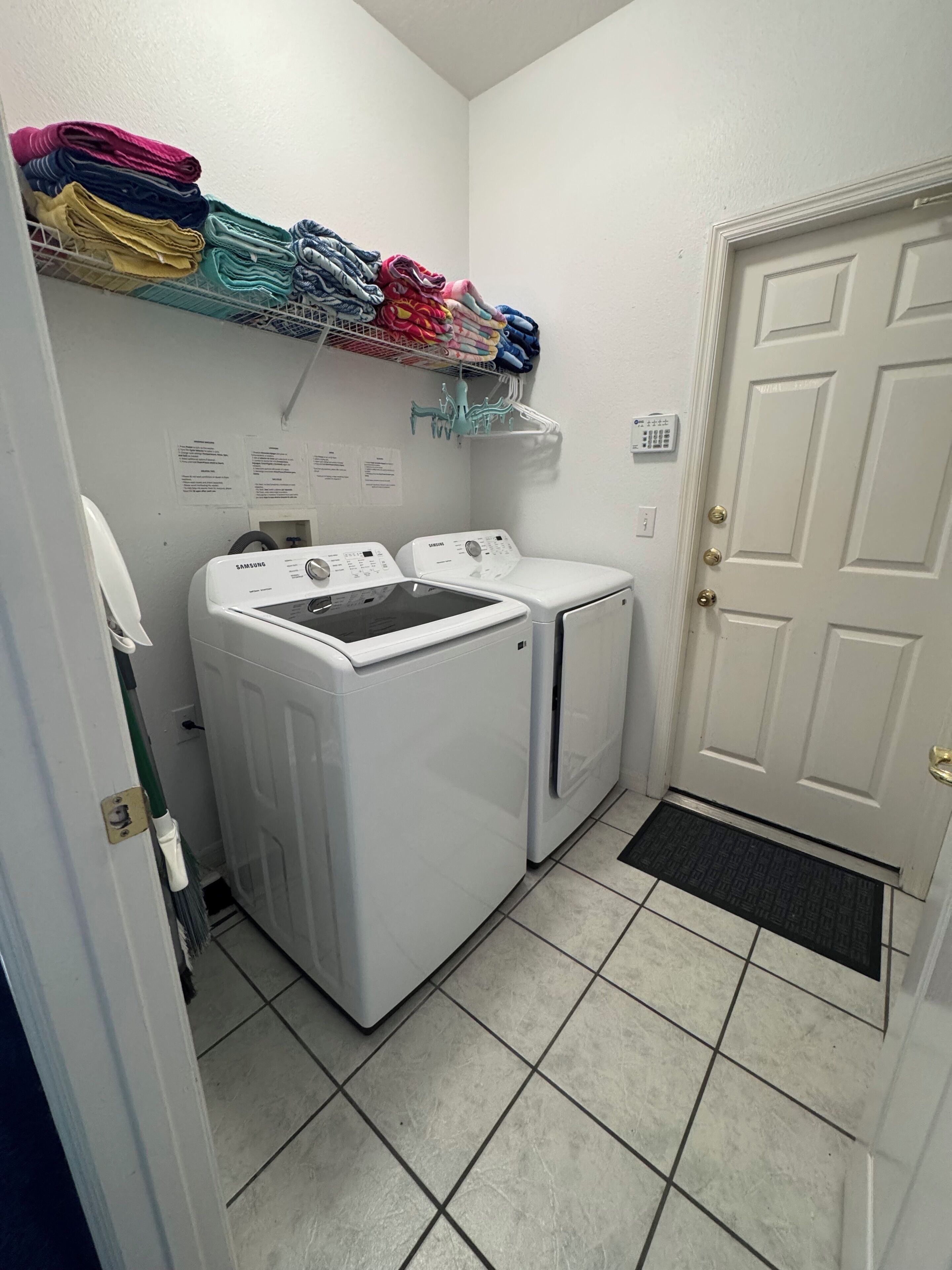 A laundry room with two white washers and dryers, a rack of folded clothes, and a door with a doormat.