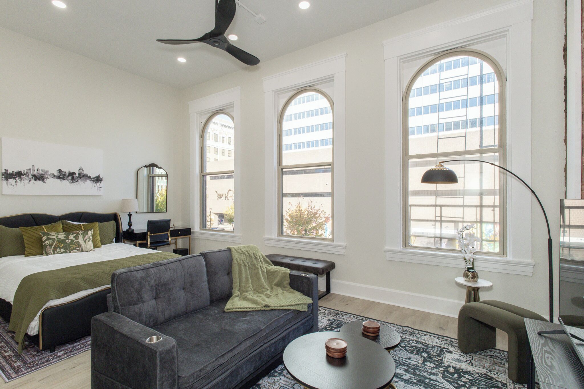 A modern hotel room with a large bed, a gray sofa, a round coffee table, and a desk with a lamp. There are three large windows providing views of the city.