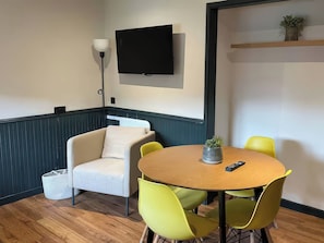 A room with a round wooden table, two yellow chairs, a white sofa, a TV mounted on the wall, a floor lamp, and a small shelf with a plant. - Ash Cottages (Llanrwst)