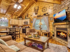 A cozy log cabin interior with a dining area, kitchen, living room, fireplace, and a television mounted on the wall.