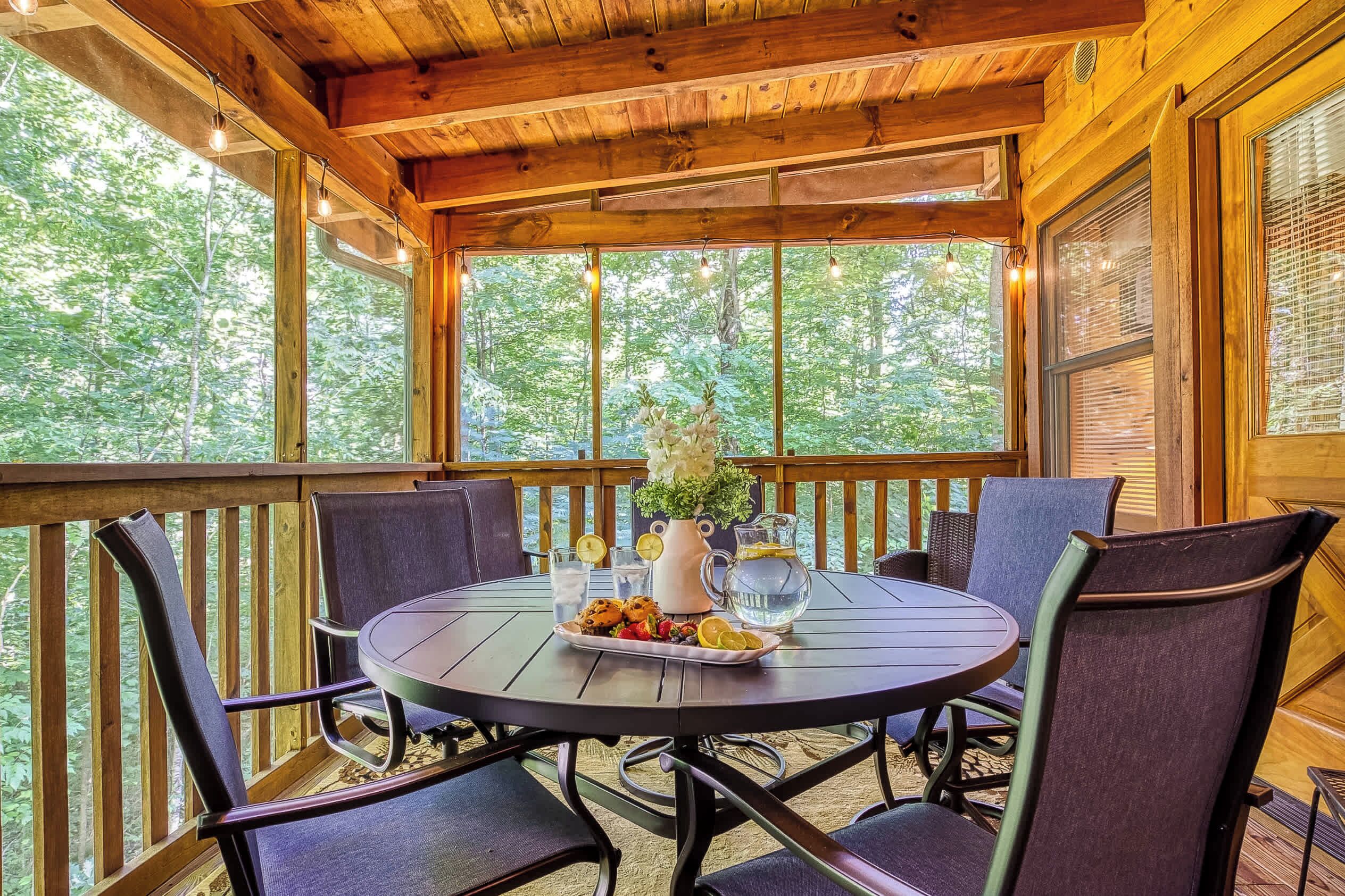 Eine Holzterrasse mit rundem Tisch und Stühlen, umgeben von großen Fenstern mit Blick auf einen Wald.