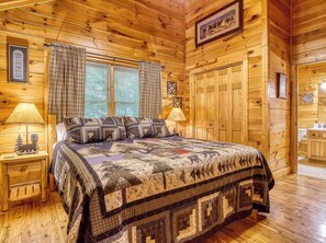 A log cabin bedroom with a large bed, two bedside lamps, plaid curtains, and wooden walls. - Quilted Pines – Hot Tub, Pool, Dog Friendly (Sevierville)