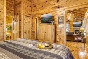 A wooden cabin interior with a bed, a television, a fireplace, and a view of the outdoors. - Quilted Pines – Pool, Fire Pit & Dog Friendly! (Sevierville)