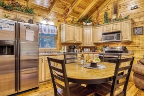 A cabin-style kitchen with wooden cabinets, a refrigerator, a microwave, and a dining table with chairs. - Quilted Pines – Hot Tub, Pool, Dog Friendly (Sevierville)