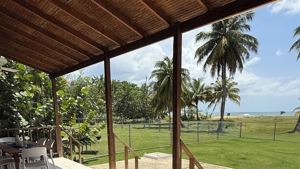 A covered outdoor seating area with white chairs and blue cushions, overlooking a beach with palm trees and a fence. - Ocean Nest - 4-bedroom beach-front house in La Playa, Anasco Puerto Rico (La Playa)