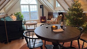 Un acogedor espacio tipo loft con techo de madera, un ventanal grande, un árbol de Navidad, una chimenea y una zona de comedor con mesa y sillas.