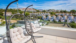 Um terraço no telhado com duas cadeiras de balanço, uma piscina e vista para uma área residencial com edifícios brancos e uma colina ao fundo.