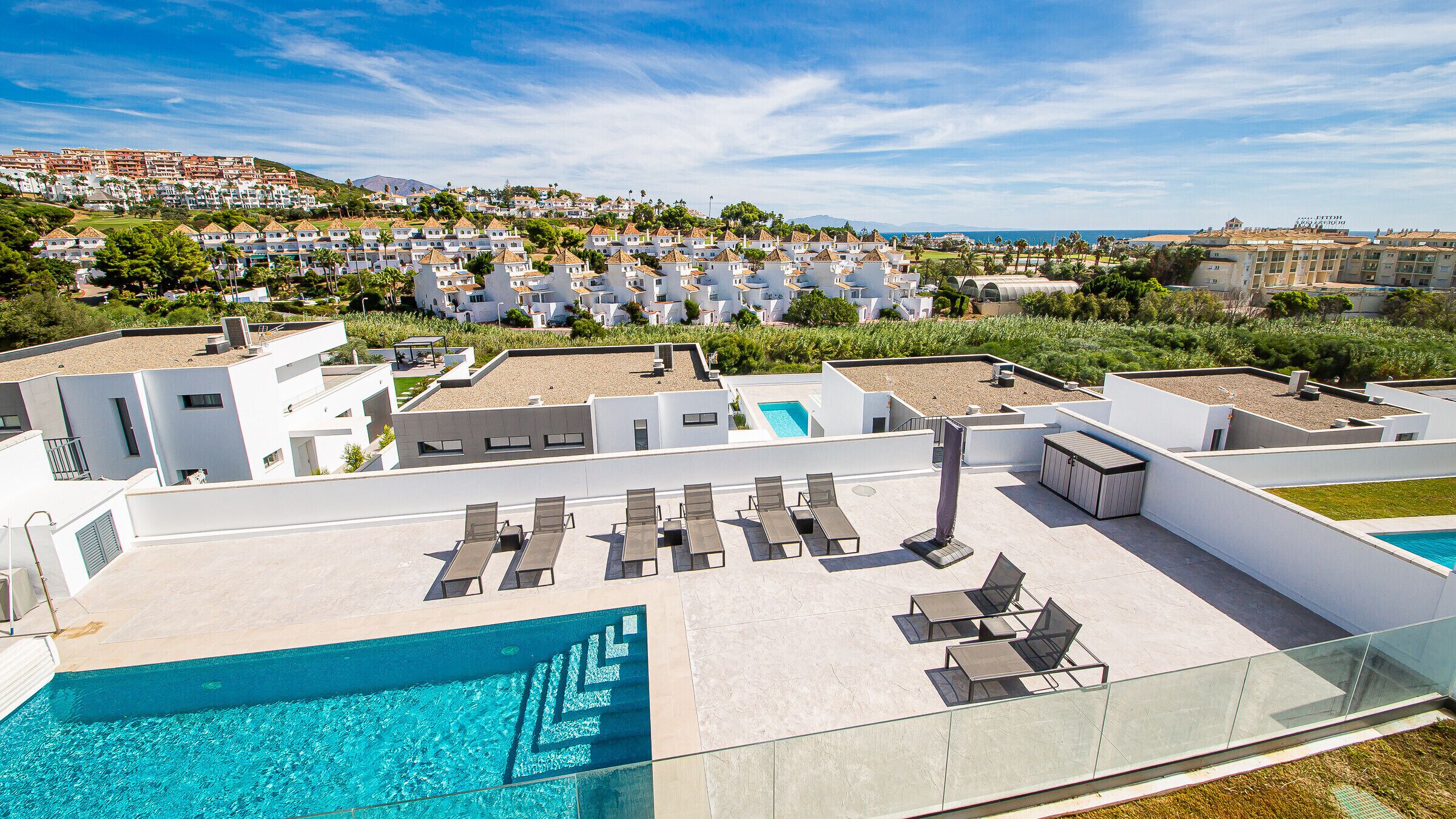 A rooftop pool area with lounge chairs and a swimming pool, surrounded by modern buildings and a view of a coastal town.